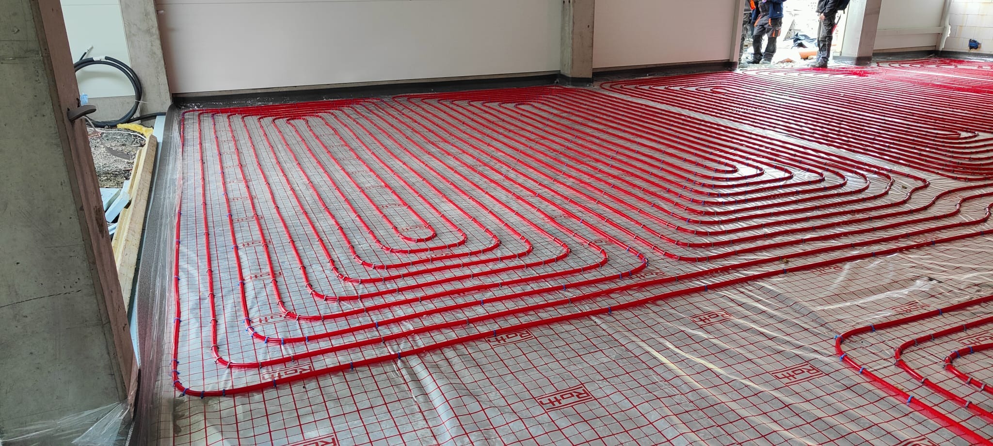Czerwone rury ogrzewania podłogowego ułożone na folii z siatką, widoczne niebieskie mocowania, surowy stan budynku, betonowe filary.