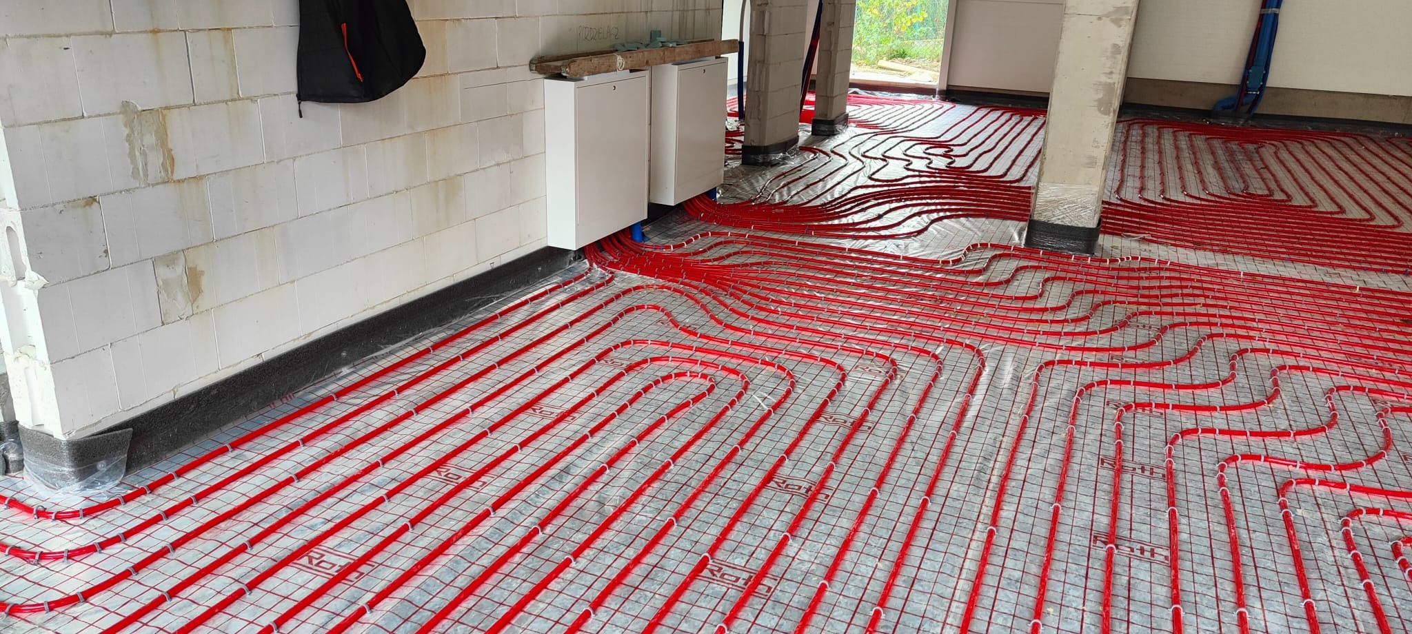 Czerwone rury ogrzewania podłogowego ułożone na srebrnej folii odblaskowej, widok z góry na nierówne podłoże w trakcie instalacji.