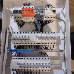 ENER-KRIS Krzysztof Bogucki - Wnętrze białej skrzynki rozdzielczej z zamontowanymi zabezpieczeniami przepięciowymi AdeliD w kolorze pomarańczowym, wyłącznikami Eaton, modułem FAEL BR4-7 i okablowaniem w kolorach czarnym...