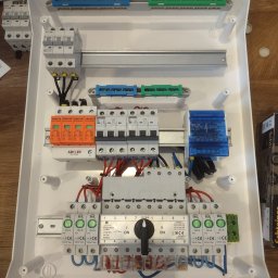 ENER-KRIS Krzysztof Bogucki - Rozdzielnica elektryczna w białej obudowie z widocznymi bezpiecznikami, wyłącznikami i przewodami, zamontowana na drewnianym podłożu, widok z góry.