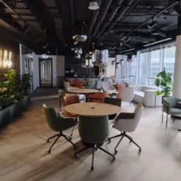 Wnętrze nowoczesnej przestrzeni biurowej z widocznymi instalacjami sufitowymi, lampami wiszącymi, stołami z krzesłami i roślinami doniczkowymi przy oknie.