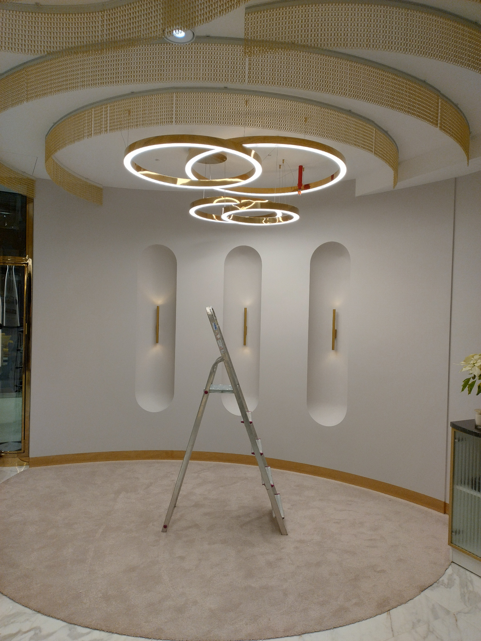 Wnętrze pomieszczenia z zainstalowanym nowoczesnym oświetleniem sufitowym w kształcie pierścieni, widoczna drabina aluminiowa, dekoracyjne oświetlenie ścienne w niszach oraz złote elementy...