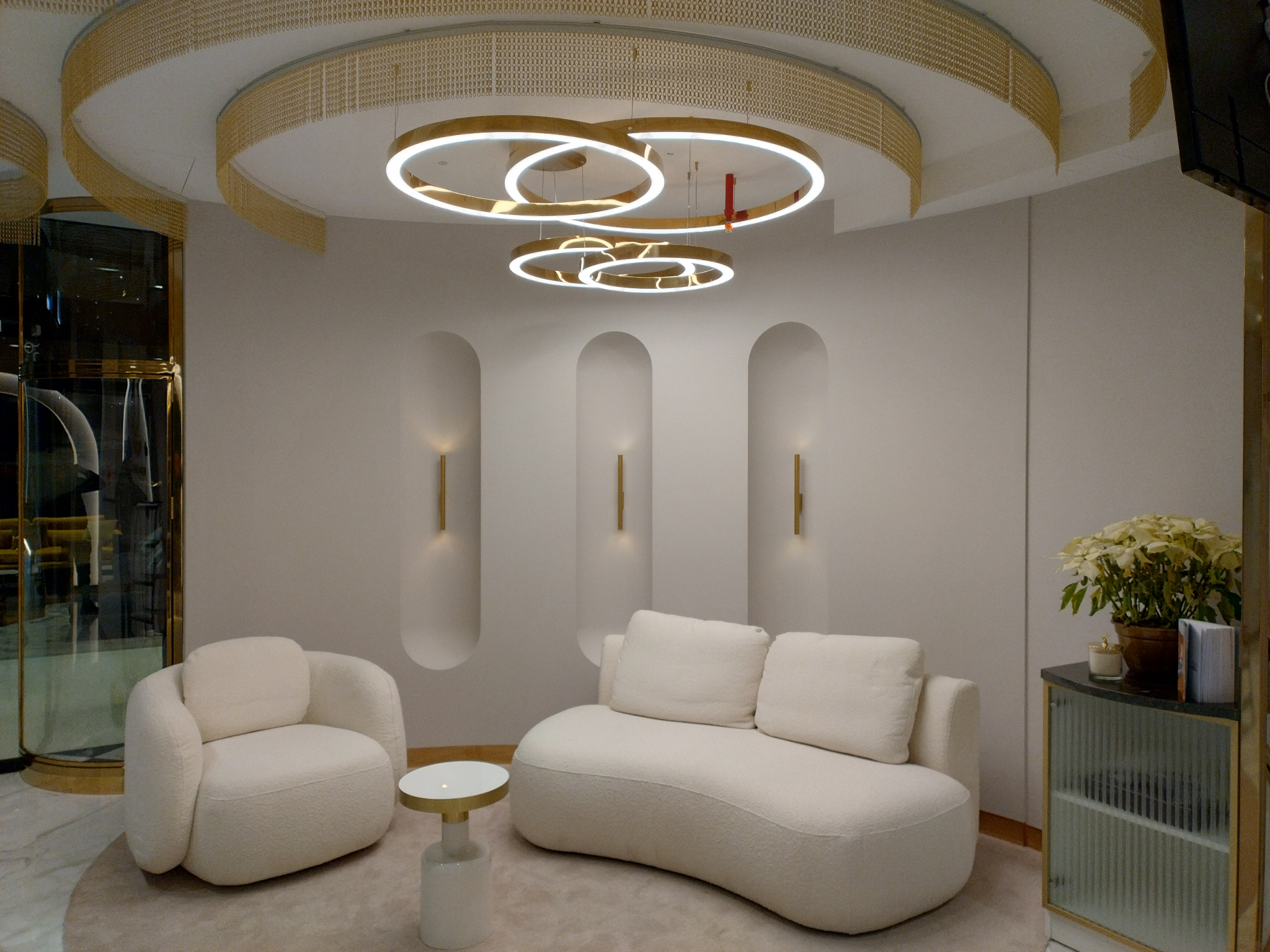 Jasne, minimalistyczne wnętrze z designerskim oświetleniem: okrągłe lampy wiszące i ścienne kinkiety. W tle kremowe meble wypoczynkowe i dekoracje.