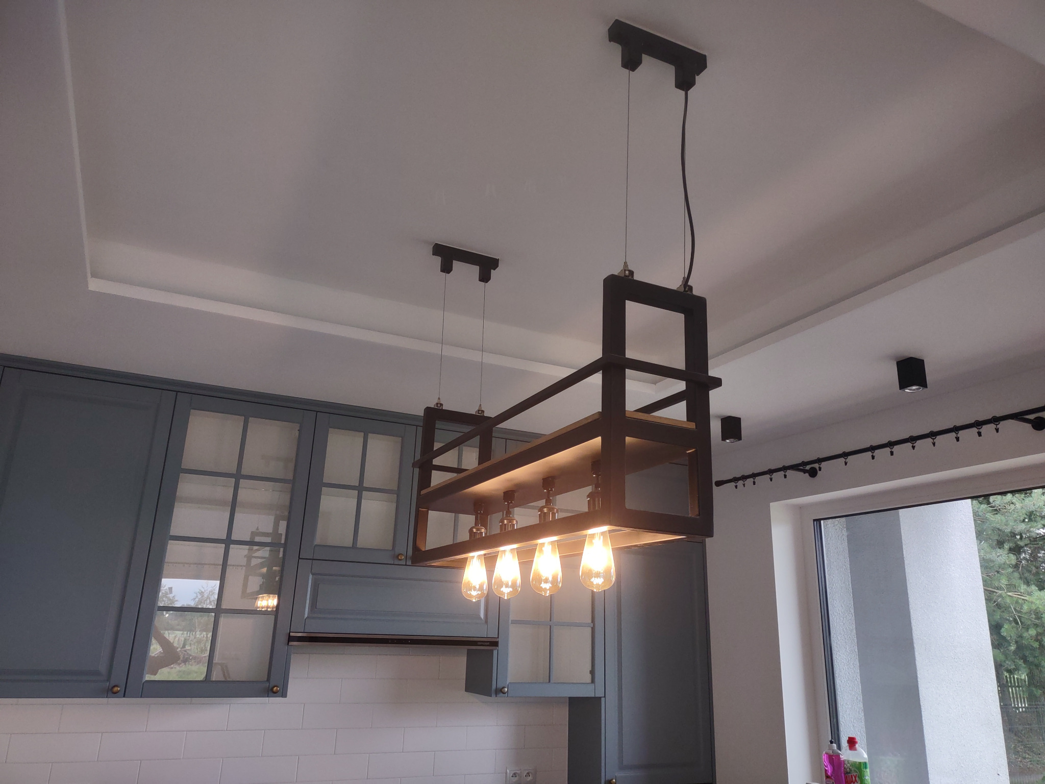 Nowoczesna lampa wisząca z czterema żarówkami Edisona, zamontowana w kuchni z szarymi szafkami i białymi płytkami, widok z dołu.