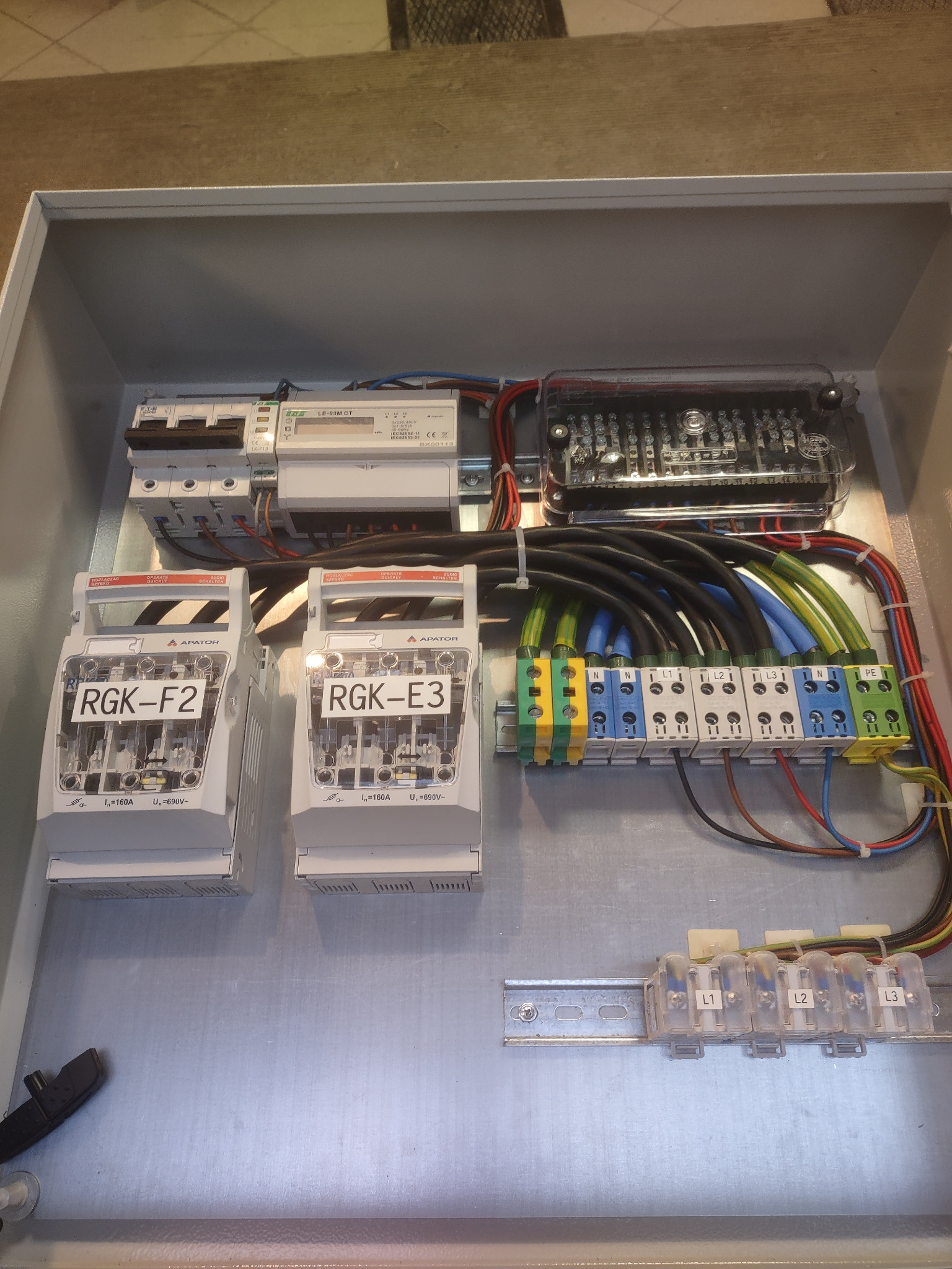 Wnętrze metalowej skrzynki rozdzielczej z zainstalowanymi bezpiecznikami RGK-F2 i RGK-E3, licznikiem energii elektrycznej LE-03M CT, listwami zaciskowymi z oznaczonymi fazami L1, L2, L3, N i PE...