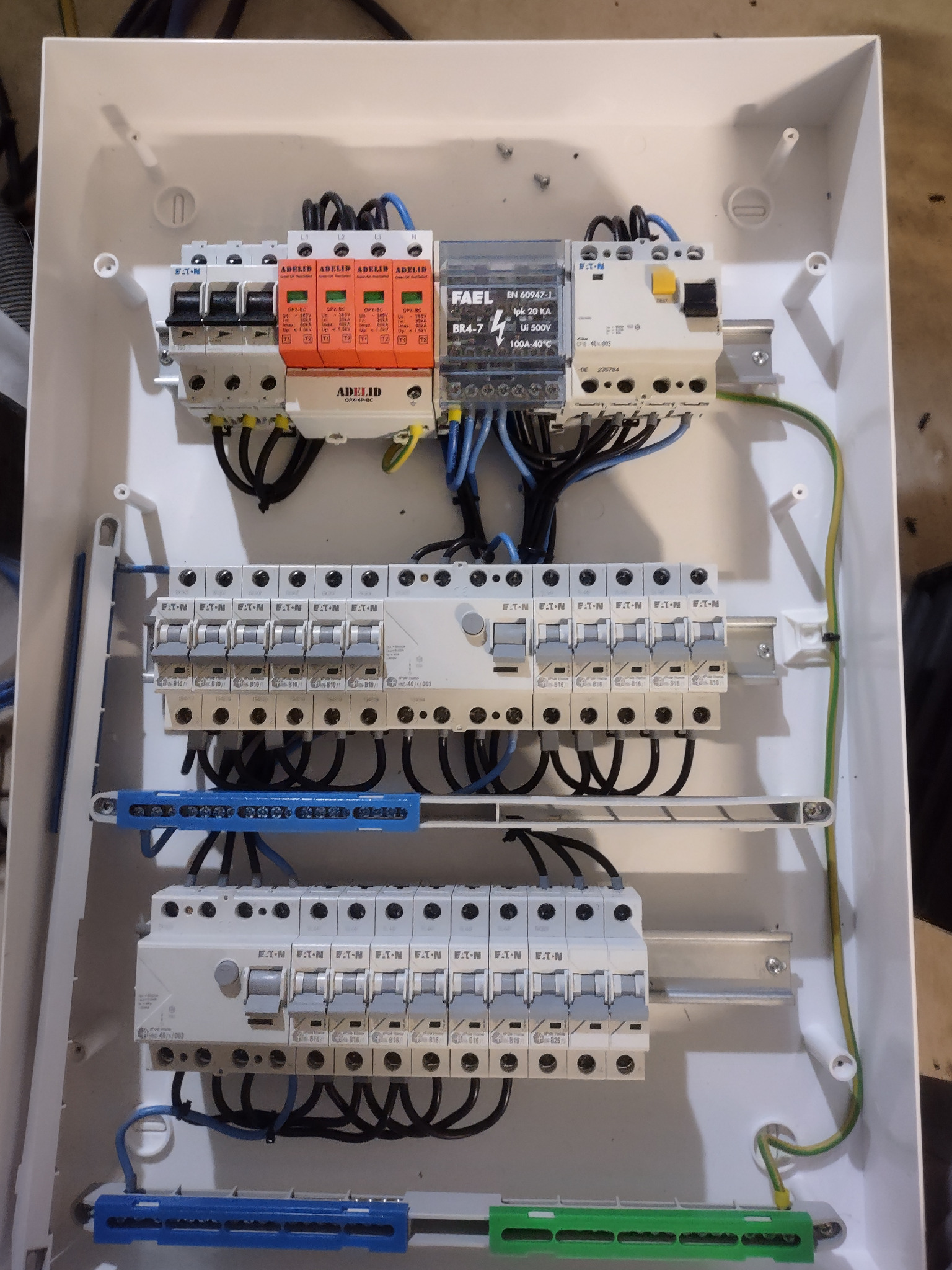 Wnętrze białej skrzynki rozdzielczej z zamontowanymi zabezpieczeniami przepięciowymi AdeliD w kolorze pomarańczowym, wyłącznikami Eaton, modułem FAEL BR4-7 i okablowaniem w kolorach czarnym...
