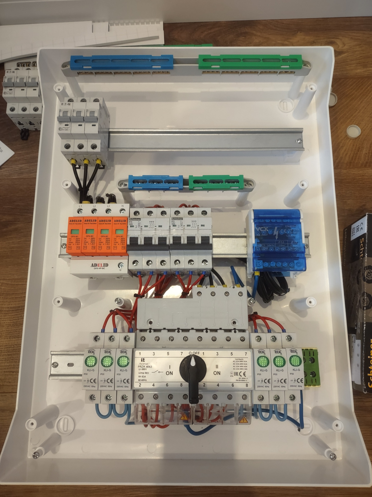 Rozdzielnica elektryczna w białej obudowie z widocznymi bezpiecznikami, wyłącznikami i przewodami, zamontowana na drewnianym podłożu, widok z góry.