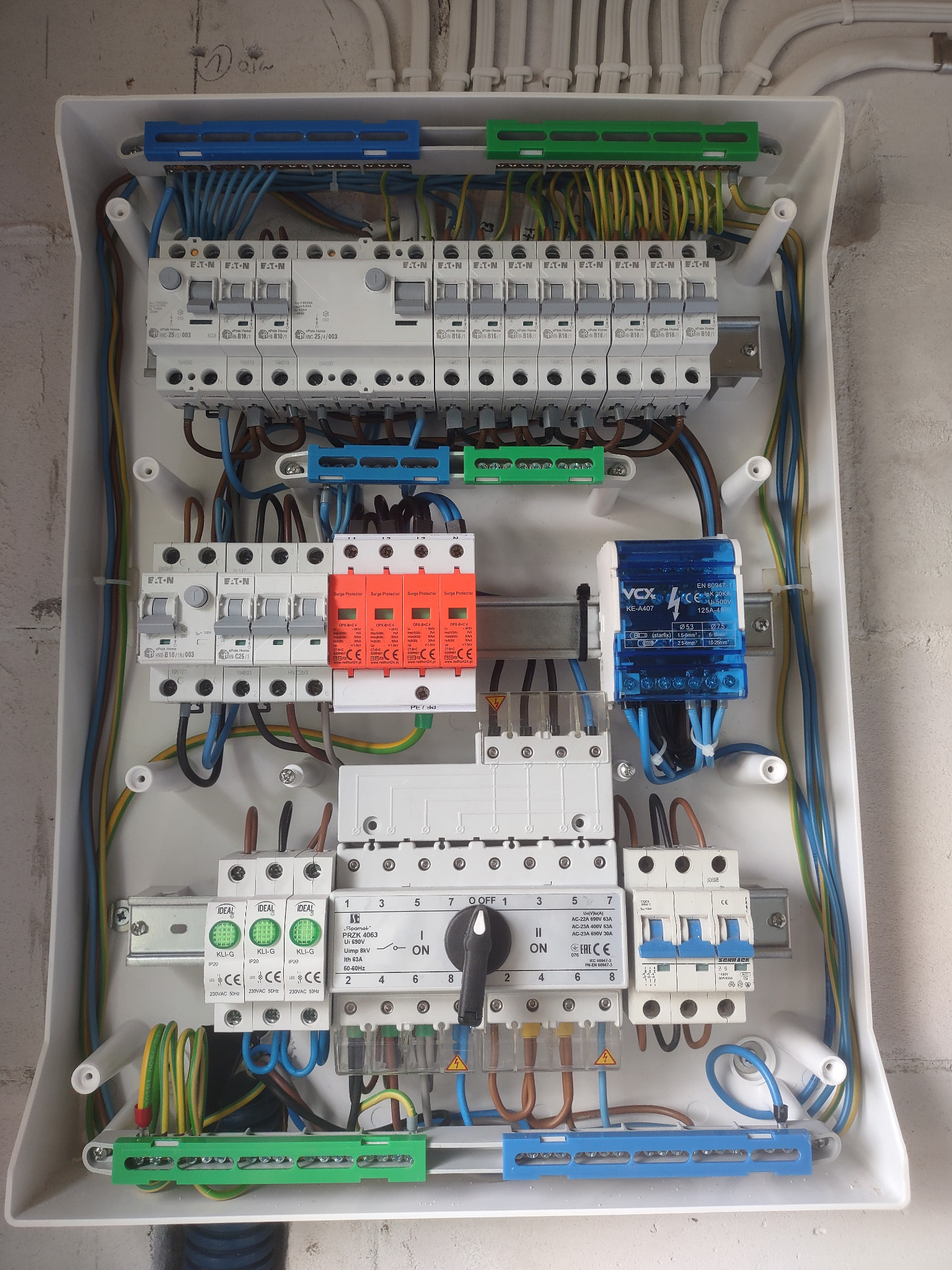 Otwarta skrzynka elektryczna z widocznymi bezpiecznikami, przewodami i innymi elementami instalacji, zamontowana na ścianie.