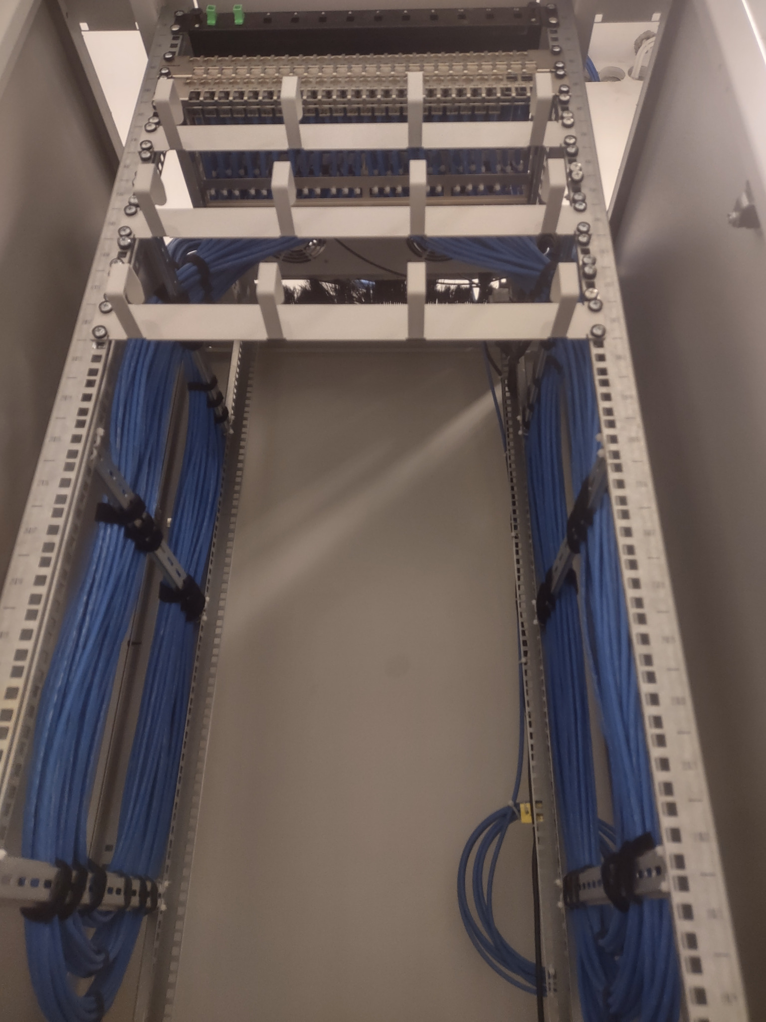 Szafa rack z uporządkowanymi niebieskimi kablami sieciowymi, widok z dołu, z widocznymi listwami zaciskowymi i elementami mocującymi kable.
