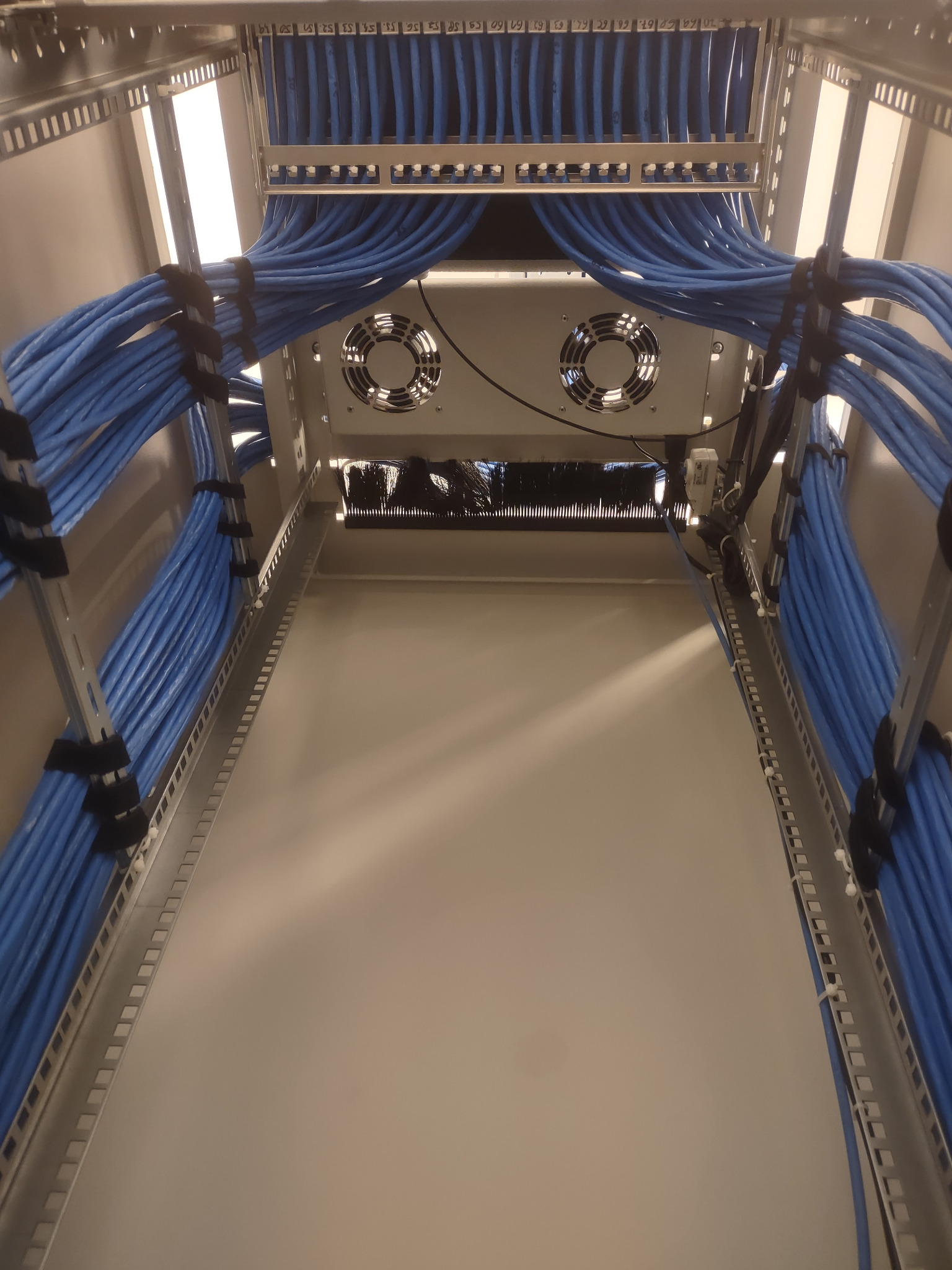 Widok z dołu szafy serwerowej z uporządkowanymi niebieskimi kablami, przymocowanymi czarnymi opaskami, oraz dwoma wentylatorami chłodzącymi na górze.