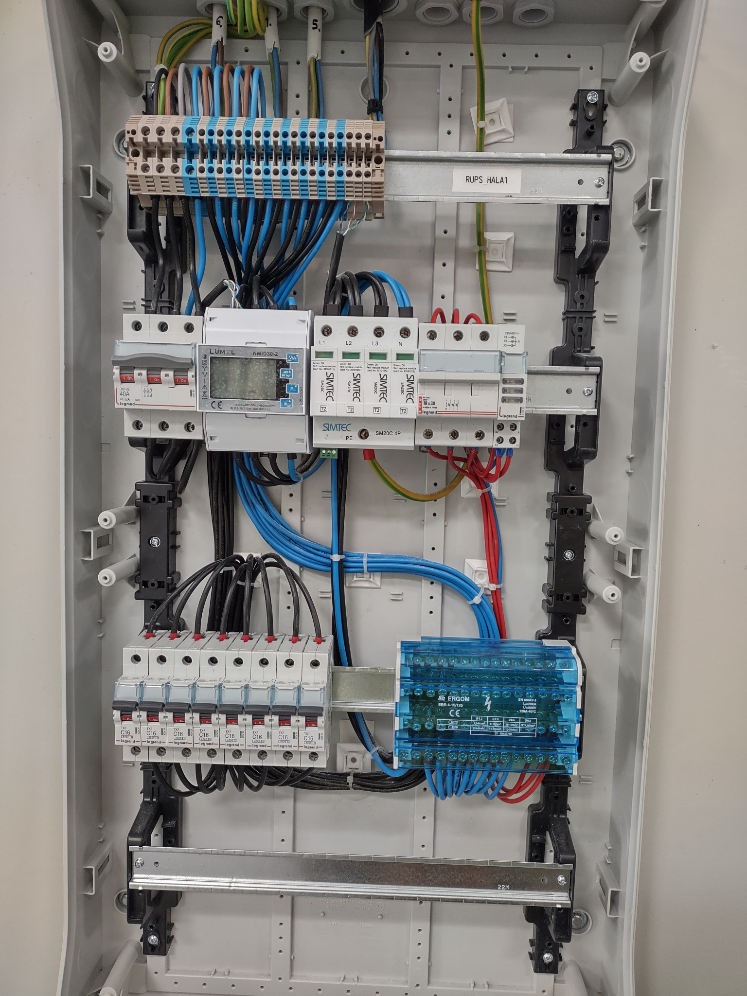 Rozdzielnica elektryczna z zamontowanymi zabezpieczeniami, licznikiem Lumel i listwami zaciskowymi, widoczne przewody w kolorach niebieskim, czarnym, czerwonym i zielono-żółtym, zamontowana...