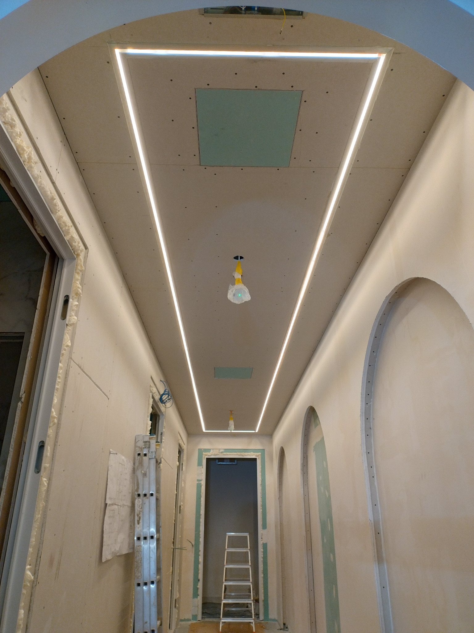 Korytarz w trakcie remontu z podwieszonym sufitem gipsowo-kartonowym, w którym zamontowano liniowe oświetlenie LED. Widoczne otwory rewizyjne, drabina i elementy wykończeniowe.