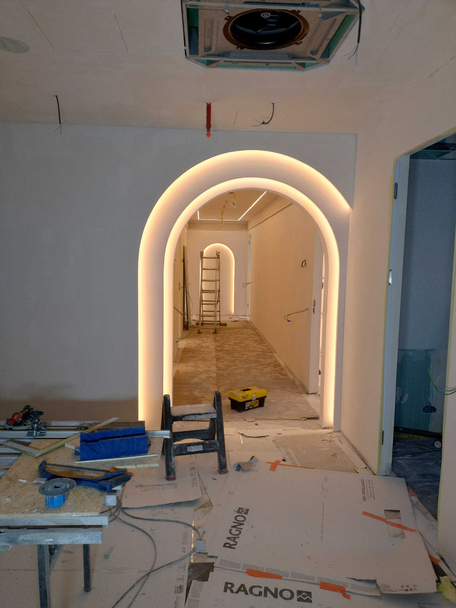 Wnętrze w trakcie wykończenia z podświetlanymi łukami, widoczne narzędzia, drabina i płyty gresowe na podłodze.