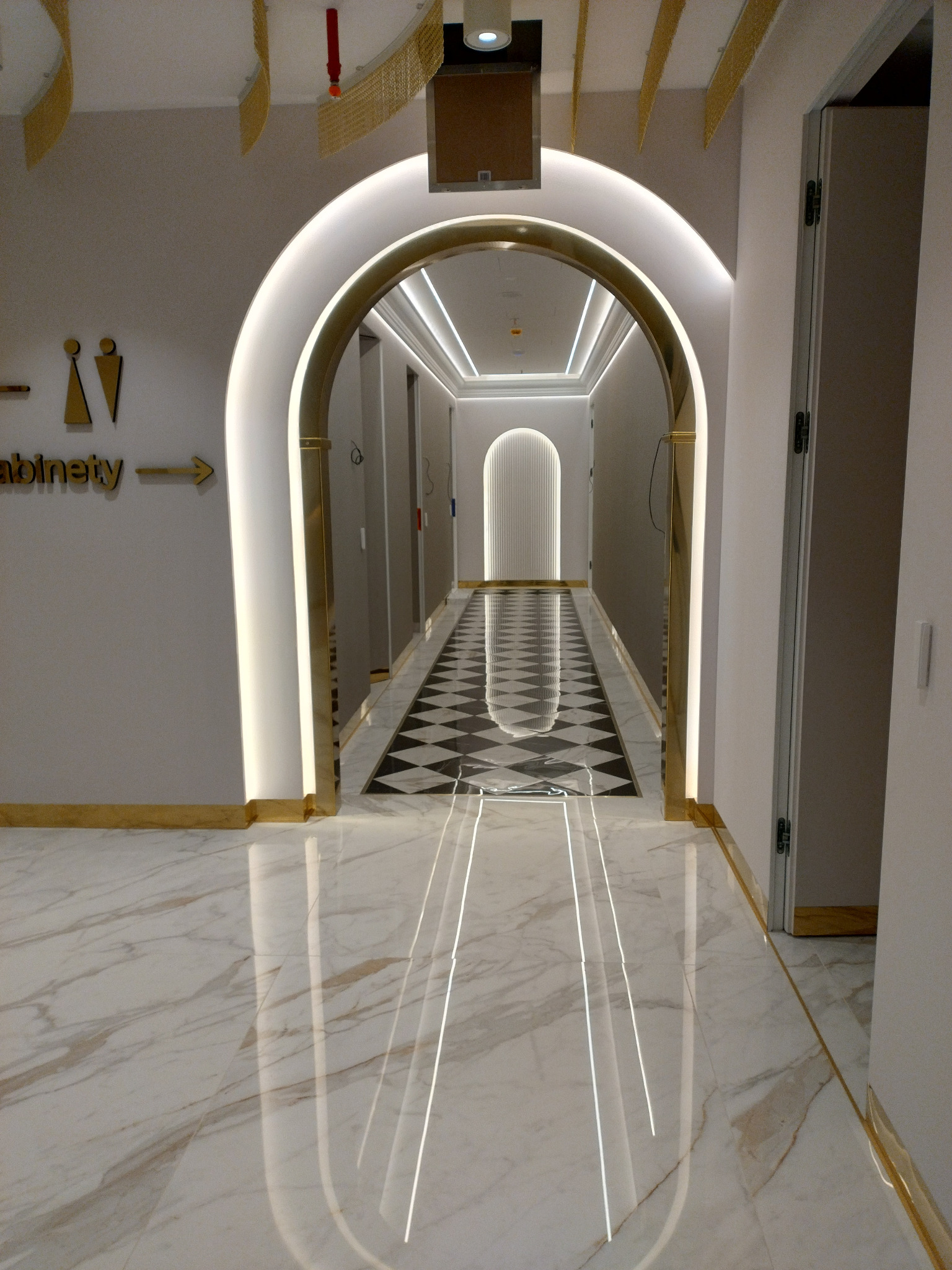 Nowoczesny korytarz z marmurową podłogą, oświetlonymi łukami i dekoracyjnymi elementami sufitu w odcieniach złota.