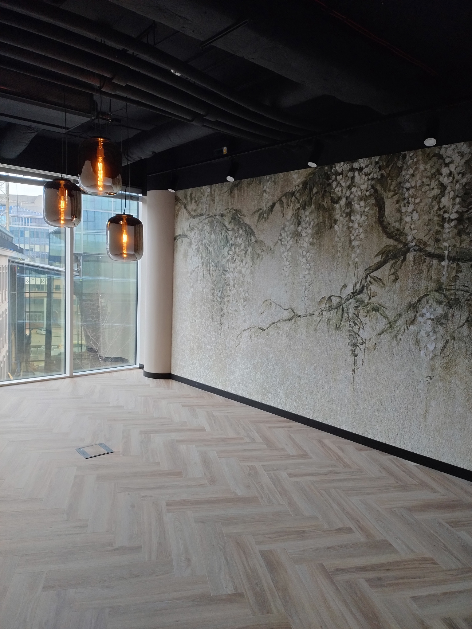 Wnętrze biurowe z drewnianą podłogą w jodełkę, ścianą z motywem roślinnym, trzema szklanymi lampami wiszącymi i widokiem na miasto za oknem, z widocznymi rurami instalacyjnymi pod sufitem.