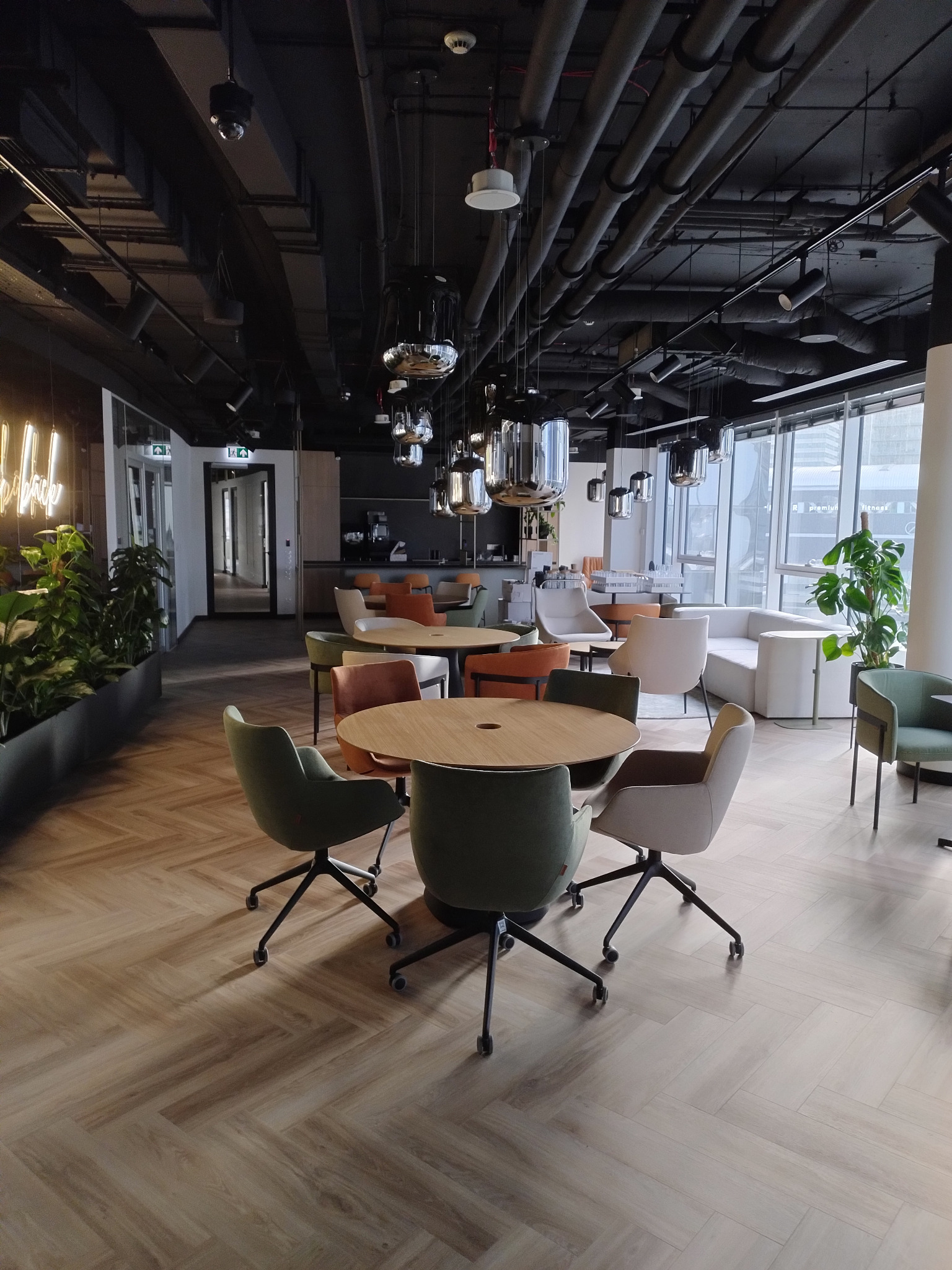 Wnętrze nowoczesnej przestrzeni biurowej z widocznymi instalacjami sufitowymi, lampami wiszącymi, stołami z krzesłami i roślinami doniczkowymi przy oknie.