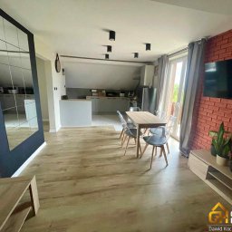 Goodhome - Jasne wnętrze po remoncie: drewniana podłoga, stół z krzesłami, ściana z cegły i TV. Widoczna kuchnia w tle. Nowoczesny design z elementami dekoracyjnymi.