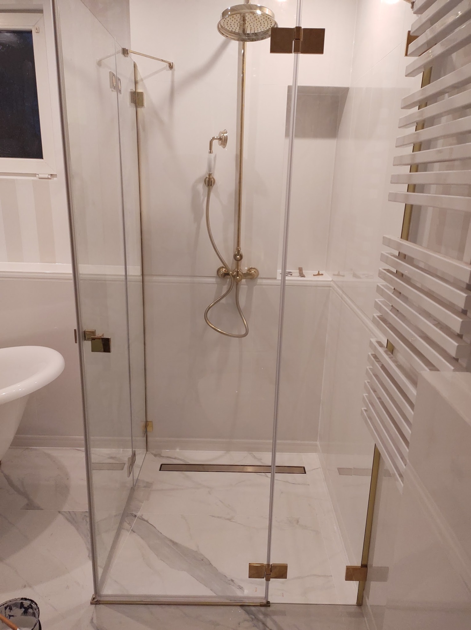 Nowoczesna kabina prysznicowa z elementami w kolorze złota, marmurowa posadzka, grzejnik drabinkowy w wykończonej łazience.