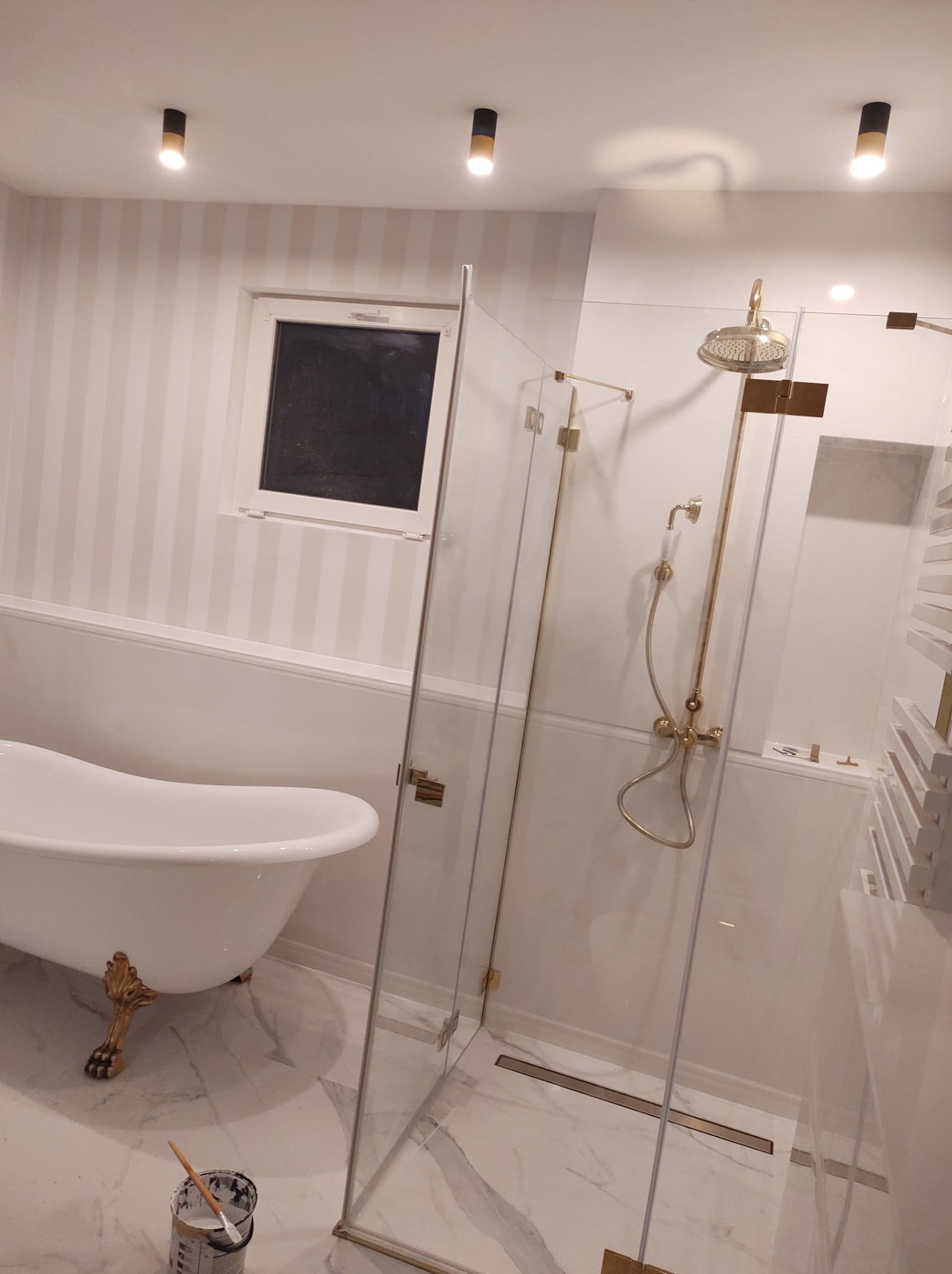 Elegancka łazienka w stylu retro z wolnostojącą wanną na złotych nóżkach, kabiną prysznicową ze złotymi okuciami i armaturą oraz ścianami w biało-szare pasy; widoczna puszka z farbą i pędzlem.