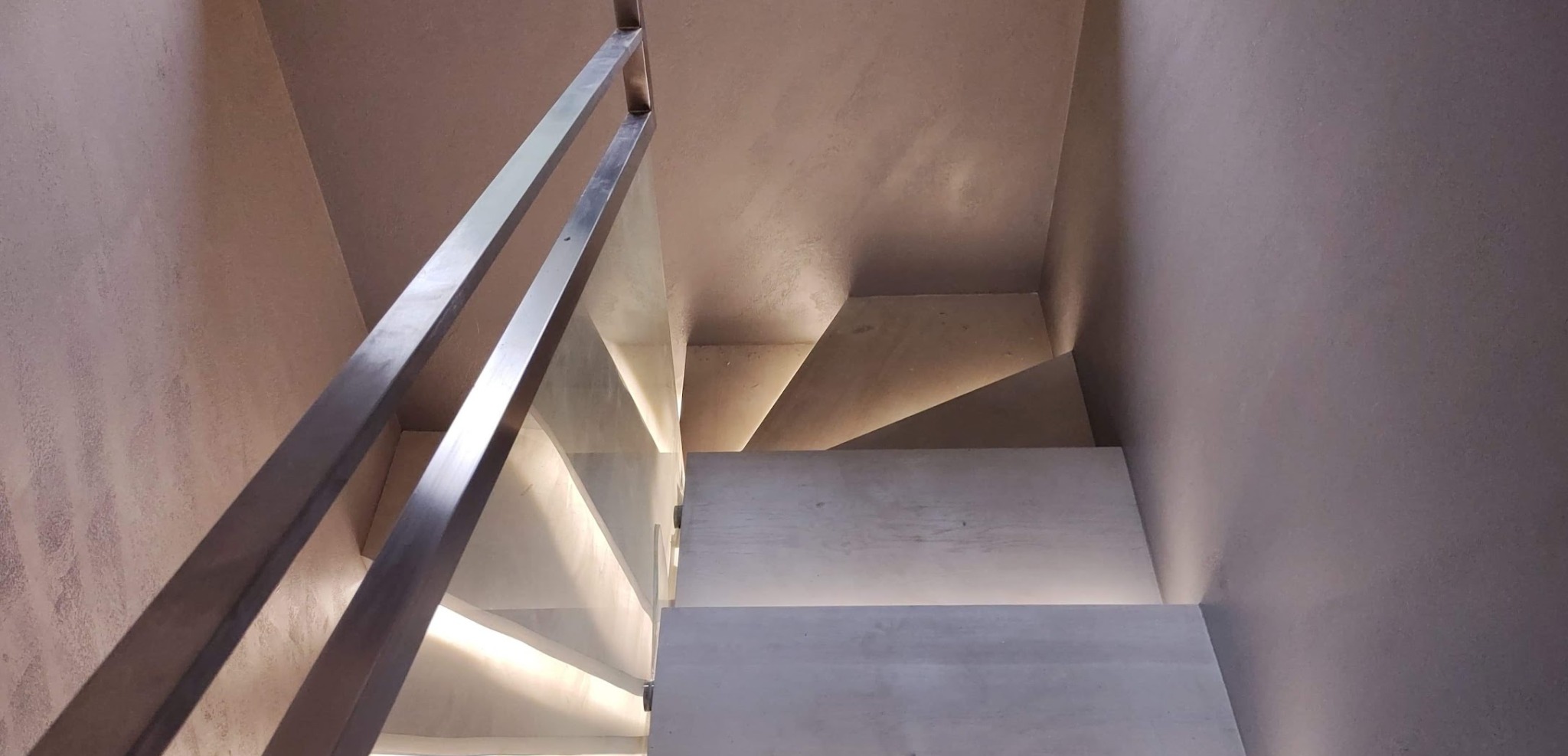 Nowoczesne schody z podświetleniem LED, metalową balustradą i szklanym wypełnieniem, ściany wykończone w odcieniach szarości.