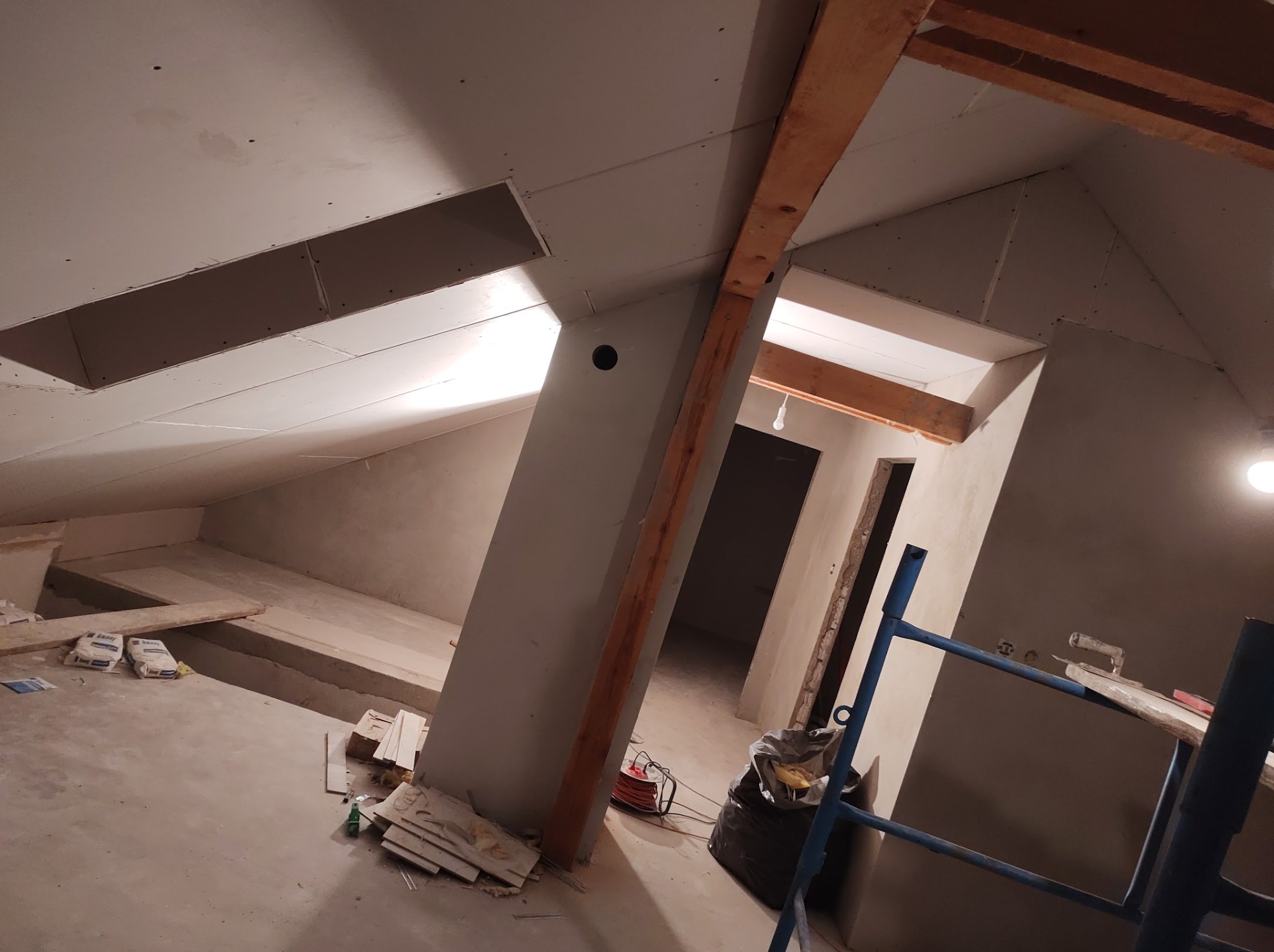 Poddasze w trakcie adaptacji: widoczne drewniane belki konstrukcyjne, zamontowane płyty kartonowo-gipsowe z otworami, częściowo wyszpachlowane ściany oraz rozstawiony rusztowanie malarskie.