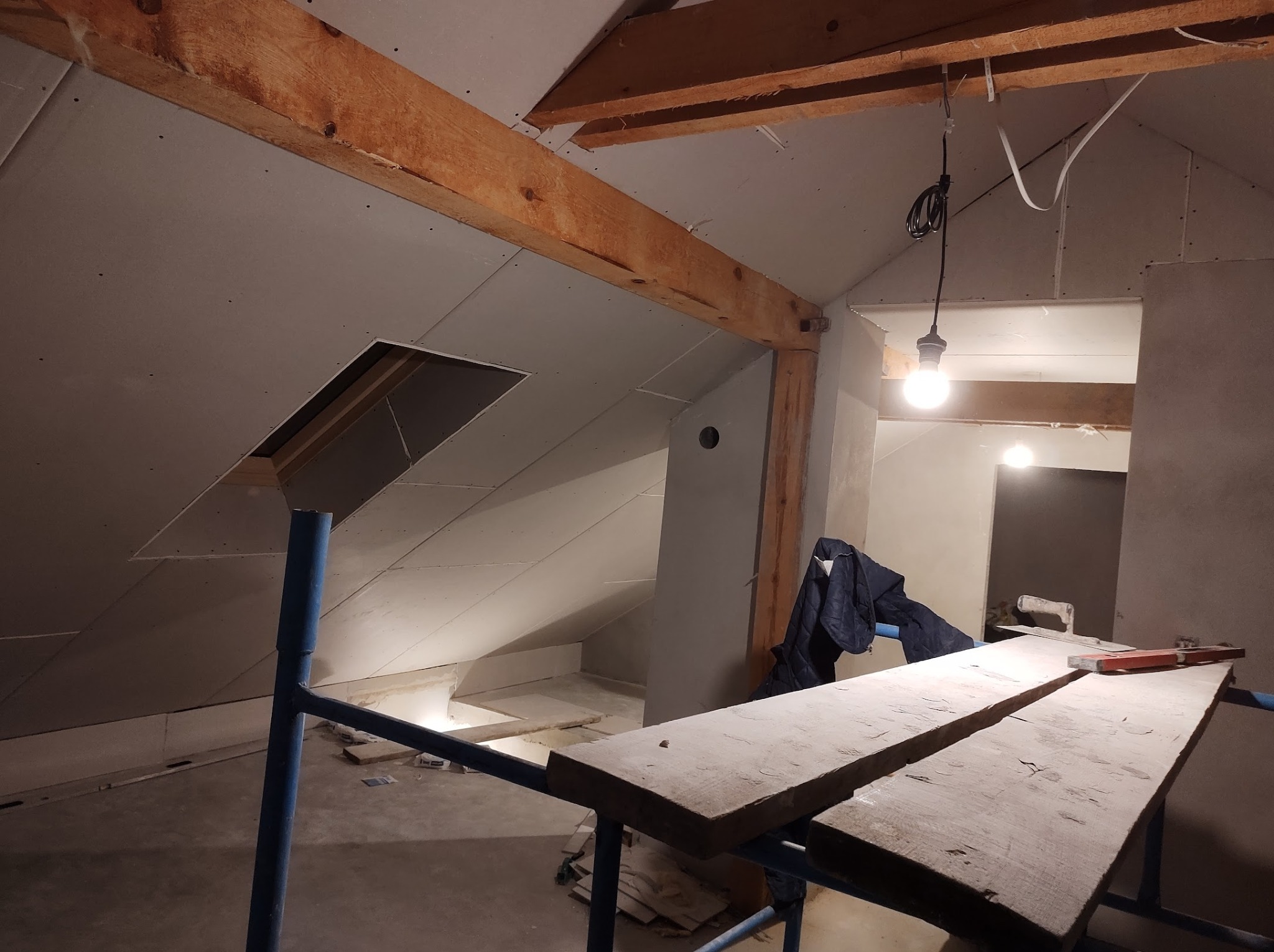 Poddasze w trakcie prac wykończeniowych: widoczne drewniane belki konstrukcyjne, zamontowane płyty karton-gips z wyciętym otworem, prowizoryczne oświetlenie żarówką oraz rusztowanie z deskami...