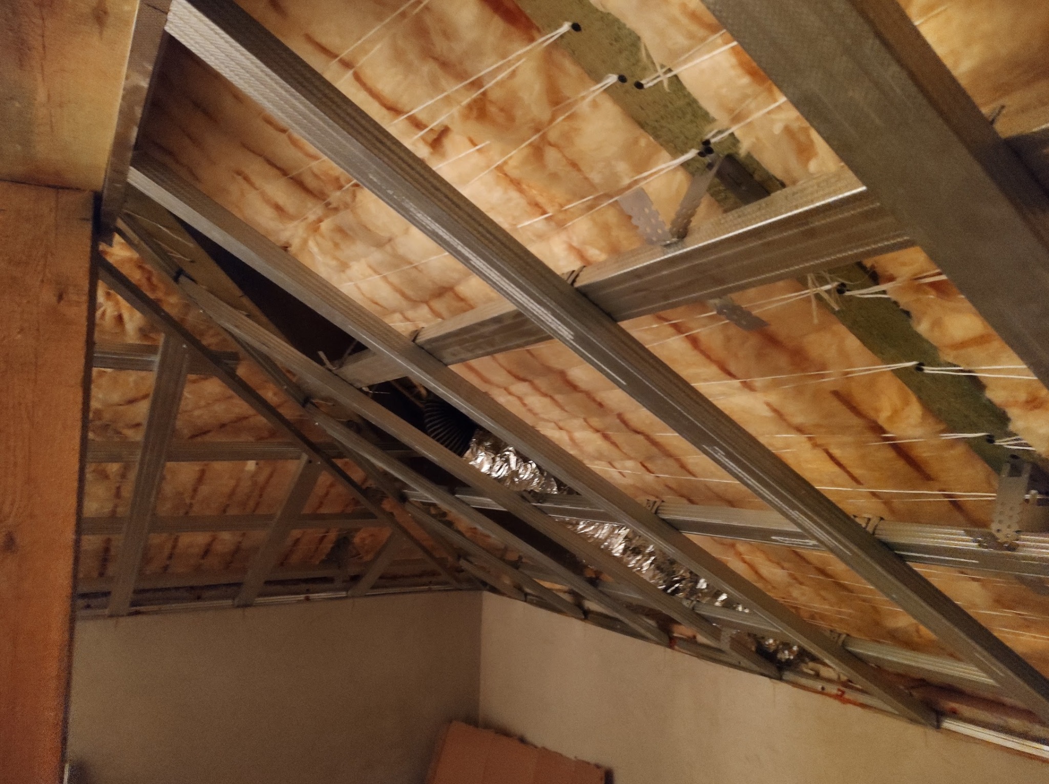 Skośny sufit poddasza z widoczną izolacją z wełny mineralnej, zamocowaną za pomocą sznurków i metalowego stelażu. Fragment wentylacji w folii aluminiowej.