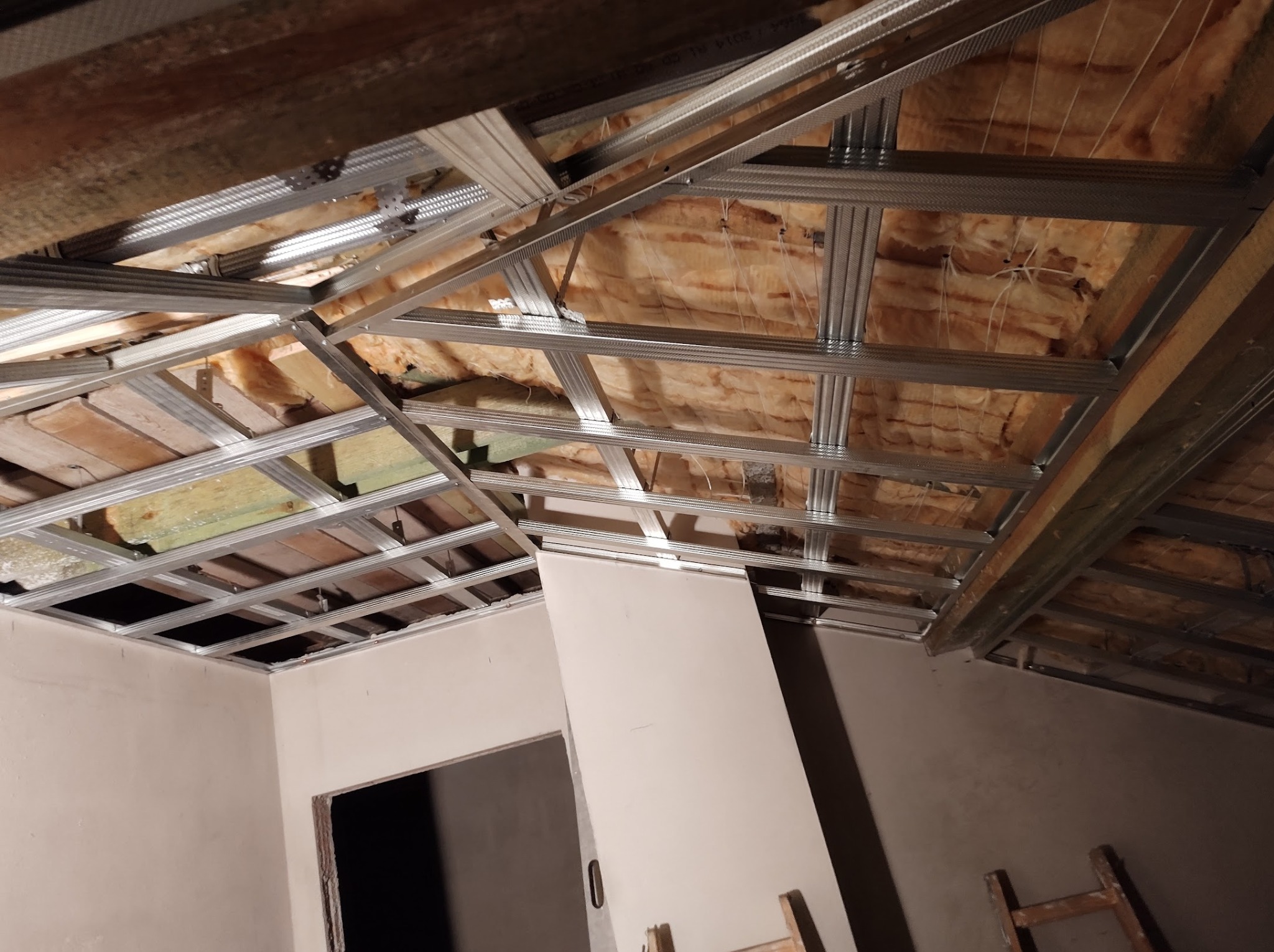 Montaż sufitu podwieszanego: metalowa konstrukcja rusztu, ocieplenie z wełny mineralnej i częściowo zamontowana płyta gipsowo-kartonowa w trakcie wykańczania poddasza.