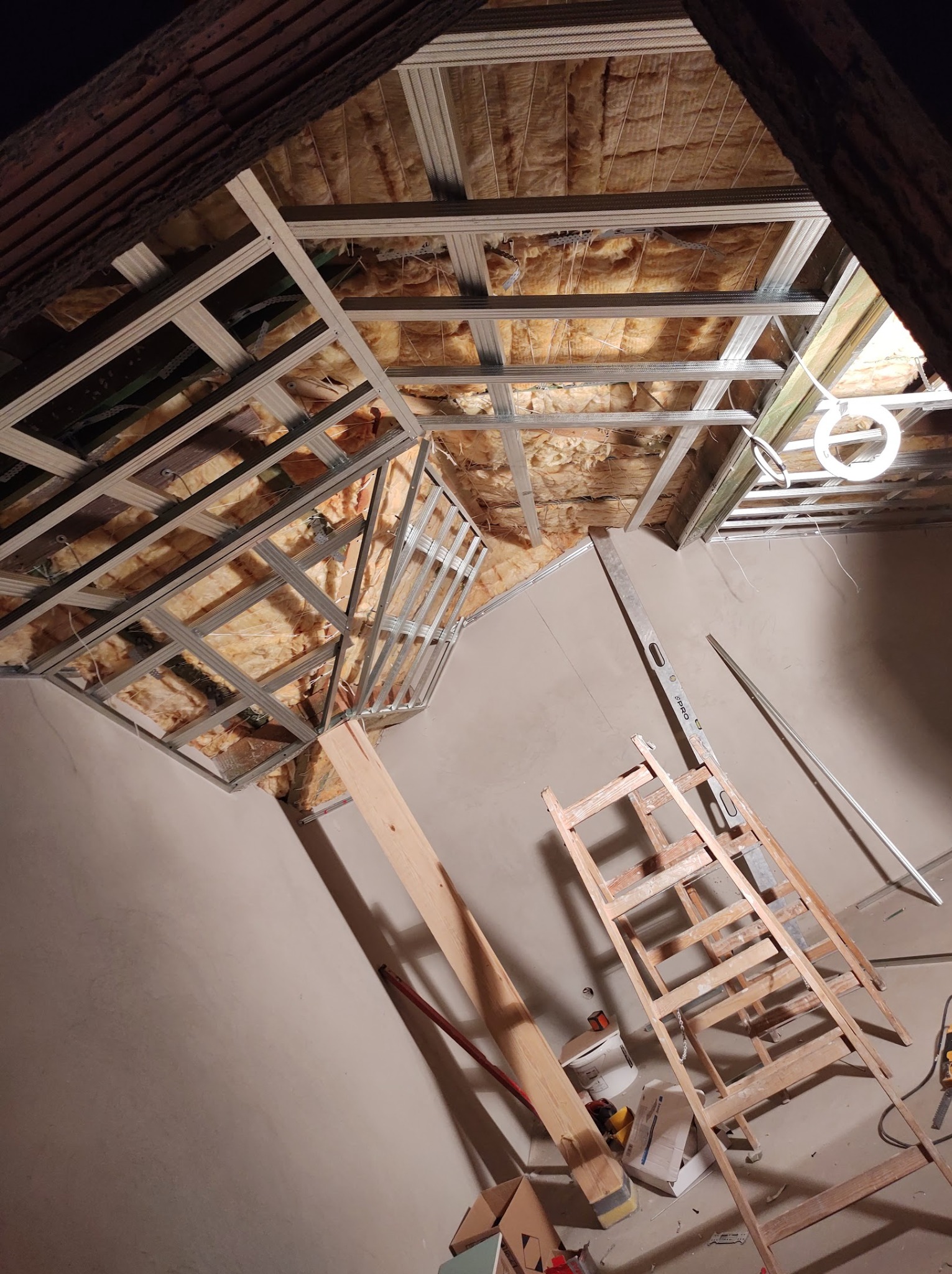Sufit poddasza w trakcie ocieplania wełną mineralną, z widoczną metalową konstrukcją pod płyty gipsowo-kartonowe, drewniana drabina i poziomica przy ścianie.