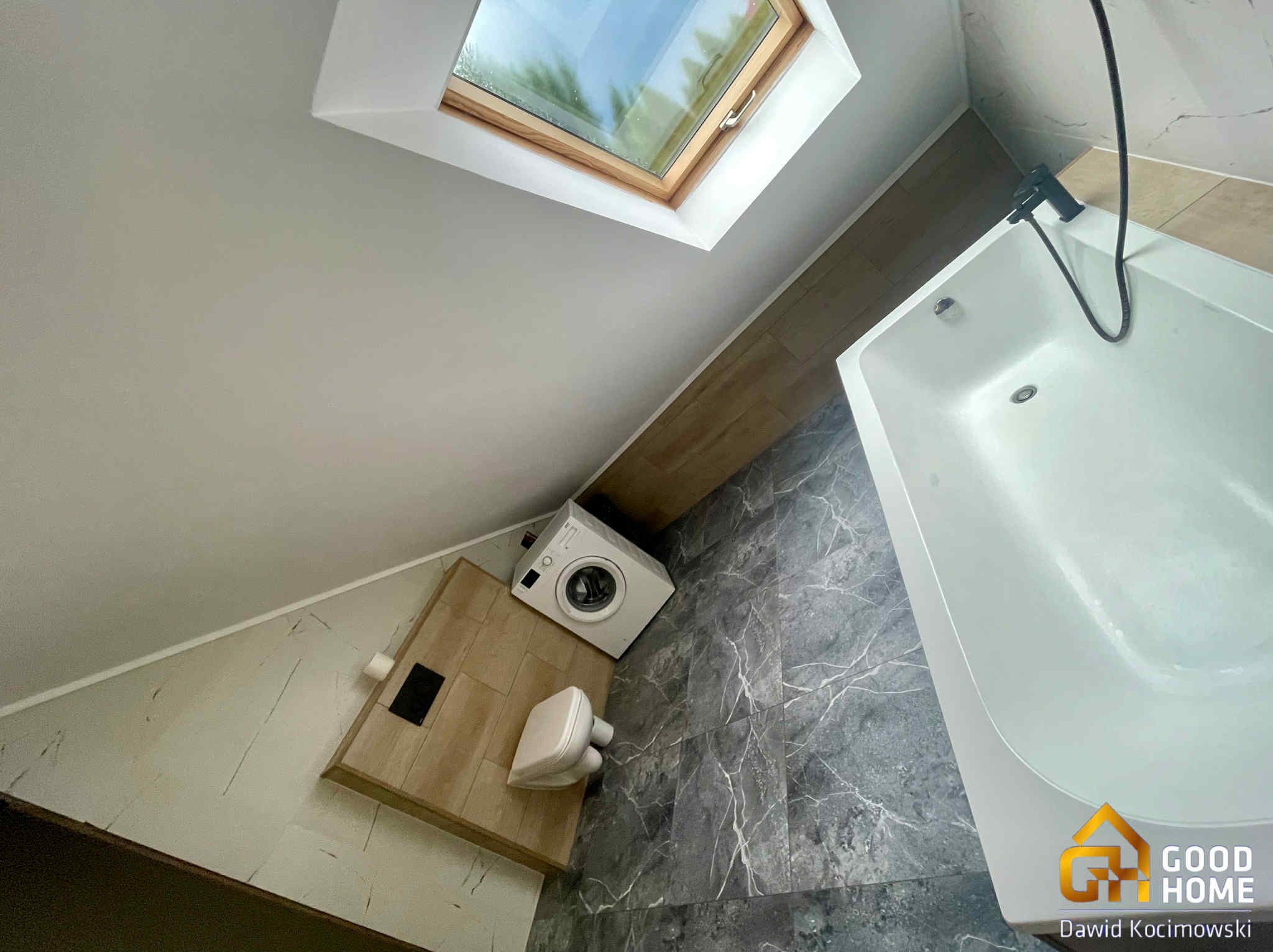 Nowoczesna łazienka na poddaszu z oknem dachowym, białą wanną i pralką. Szare płytki imitujące marmur na podłodze i ścianach. Ujęcie z góry.