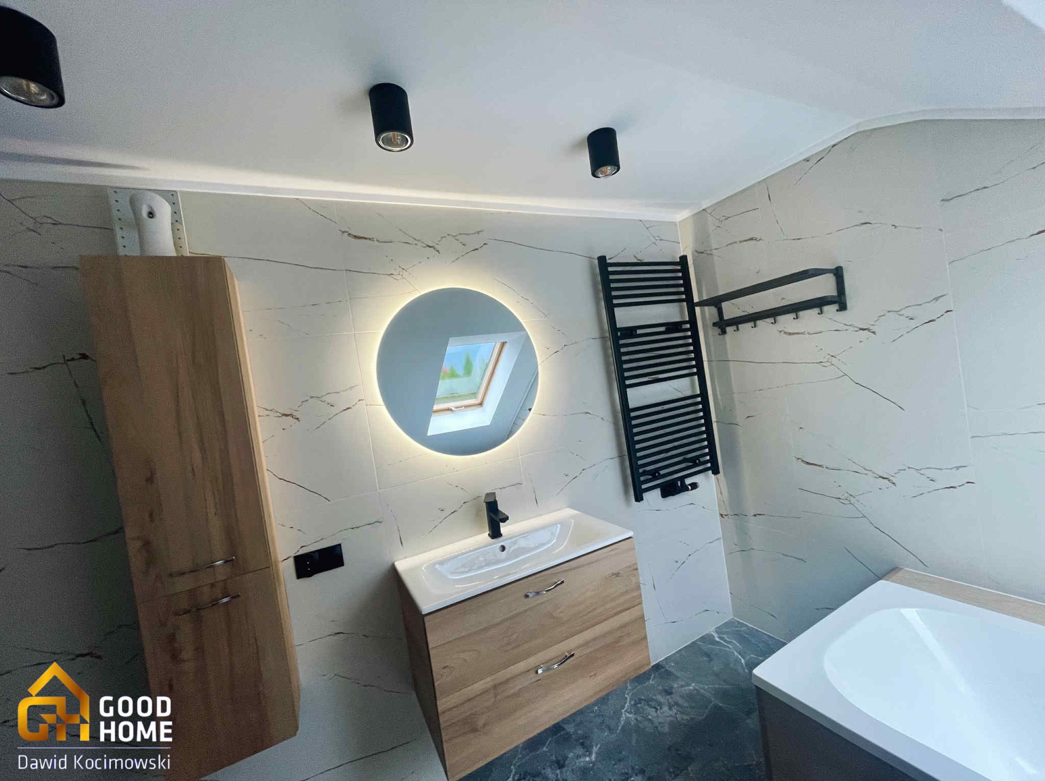 Nowoczesna łazienka z okrągłym lustrem z podświetleniem LED, drewnianą szafką pod umywalką i grzejnikiem drabinkowym na ścianie z marmurowym wzorem.