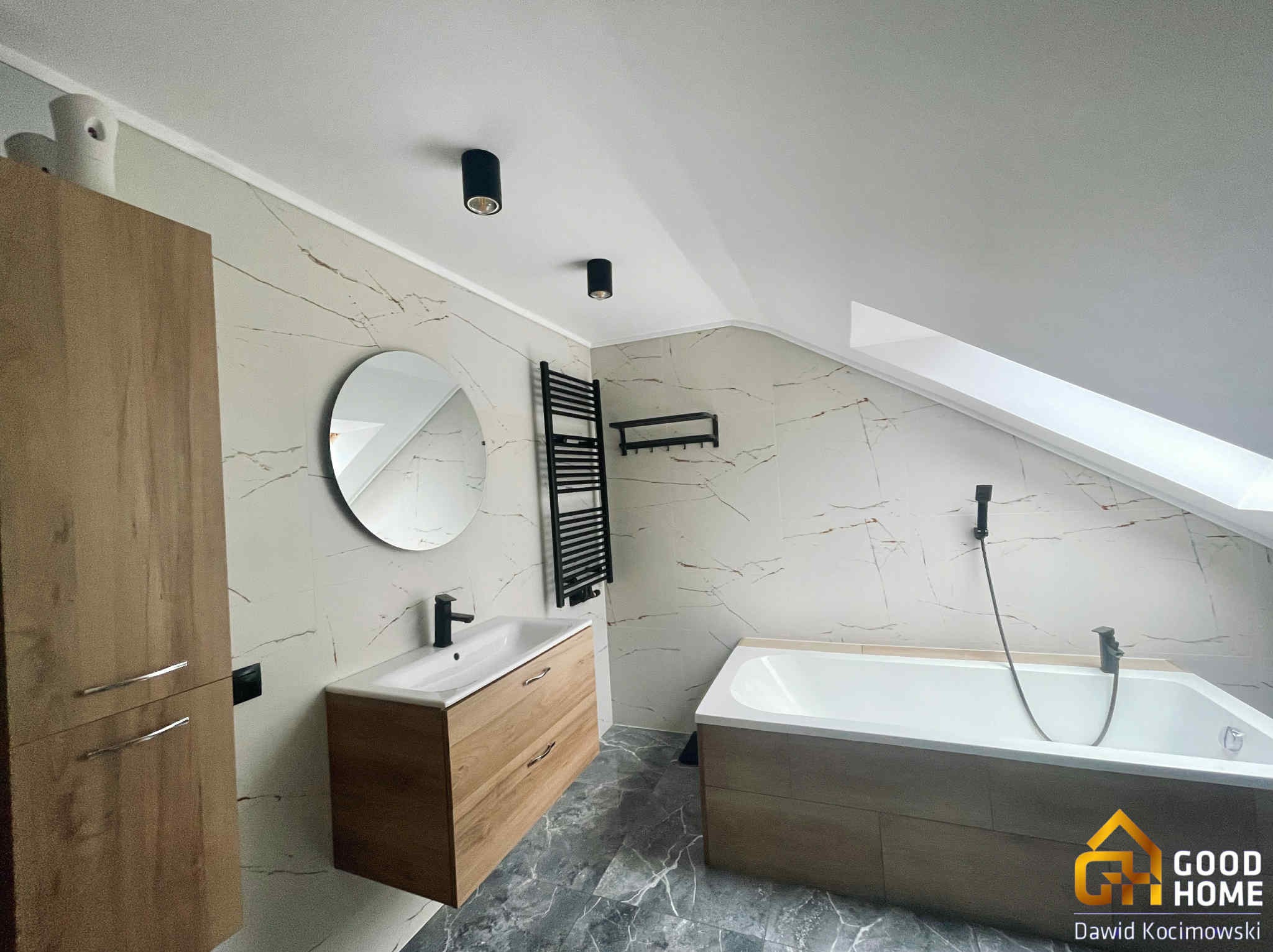 Stylowa łazienka na poddaszu z marmurowymi płytkami, drewnianymi szafkami, okrągłym lustrem i czarnymi lampami sufitowymi. Umywalka z baterią i wanna.