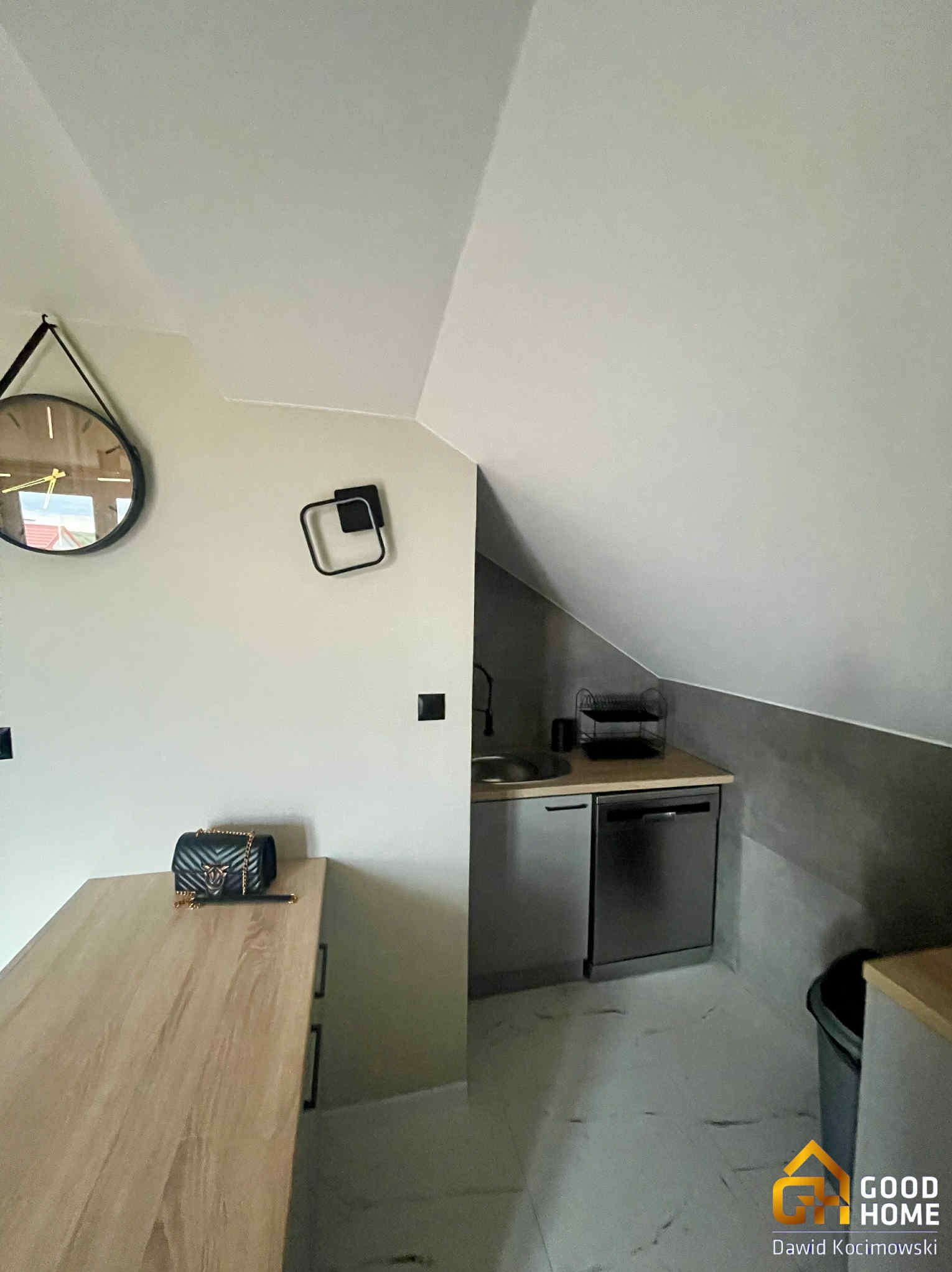 Klimatyczna kuchnia na poddaszu ze skośnym sufitem. Widoczny zlew, szafki, marmurowe płytki podłogowe, dekoracje na ścianie i torebka na blacie.