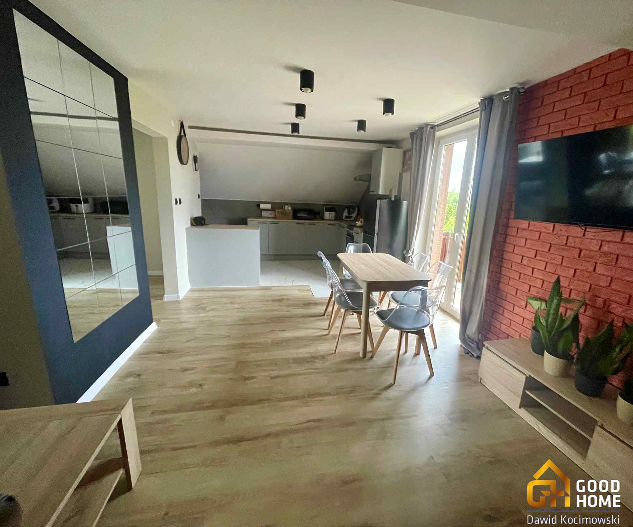 Jasne wnętrze po remoncie: drewniana podłoga, stół z krzesłami, ściana z cegły i TV. Widoczna kuchnia w tle. Nowoczesny design z elementami dekoracyjnymi.