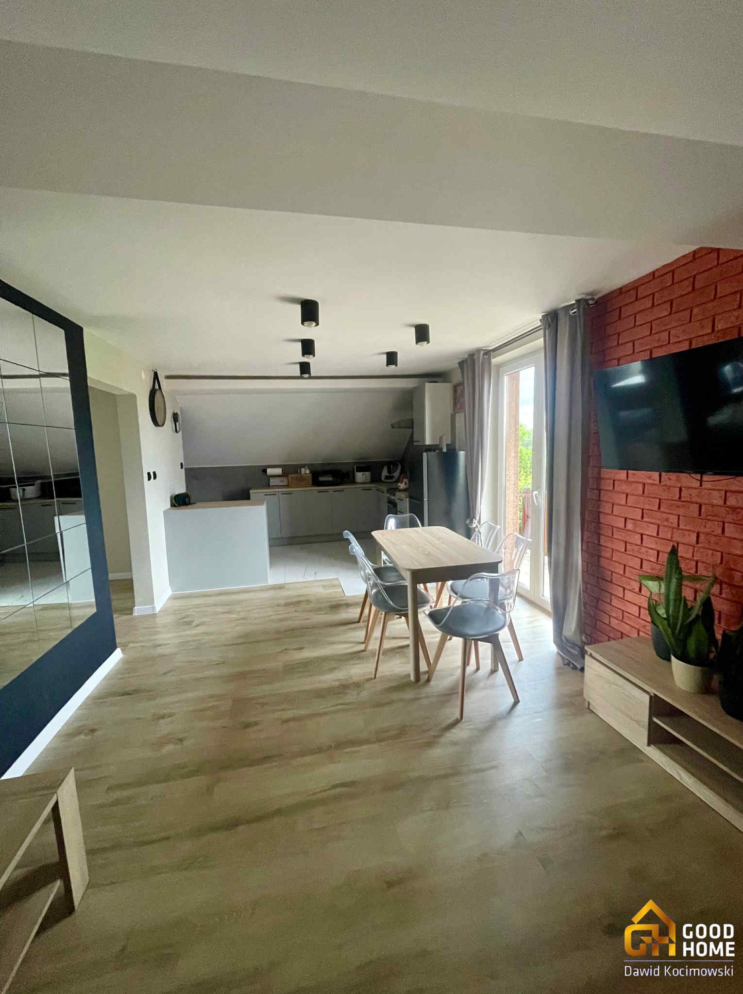 Przestronne wnętrze: parkiet, stół z krzesłami, ceglana ściana z TV, lustro w czarnej ramie. Widok na kuchnię. Nowoczesny design, jasne kolory, lampy sufitowe.