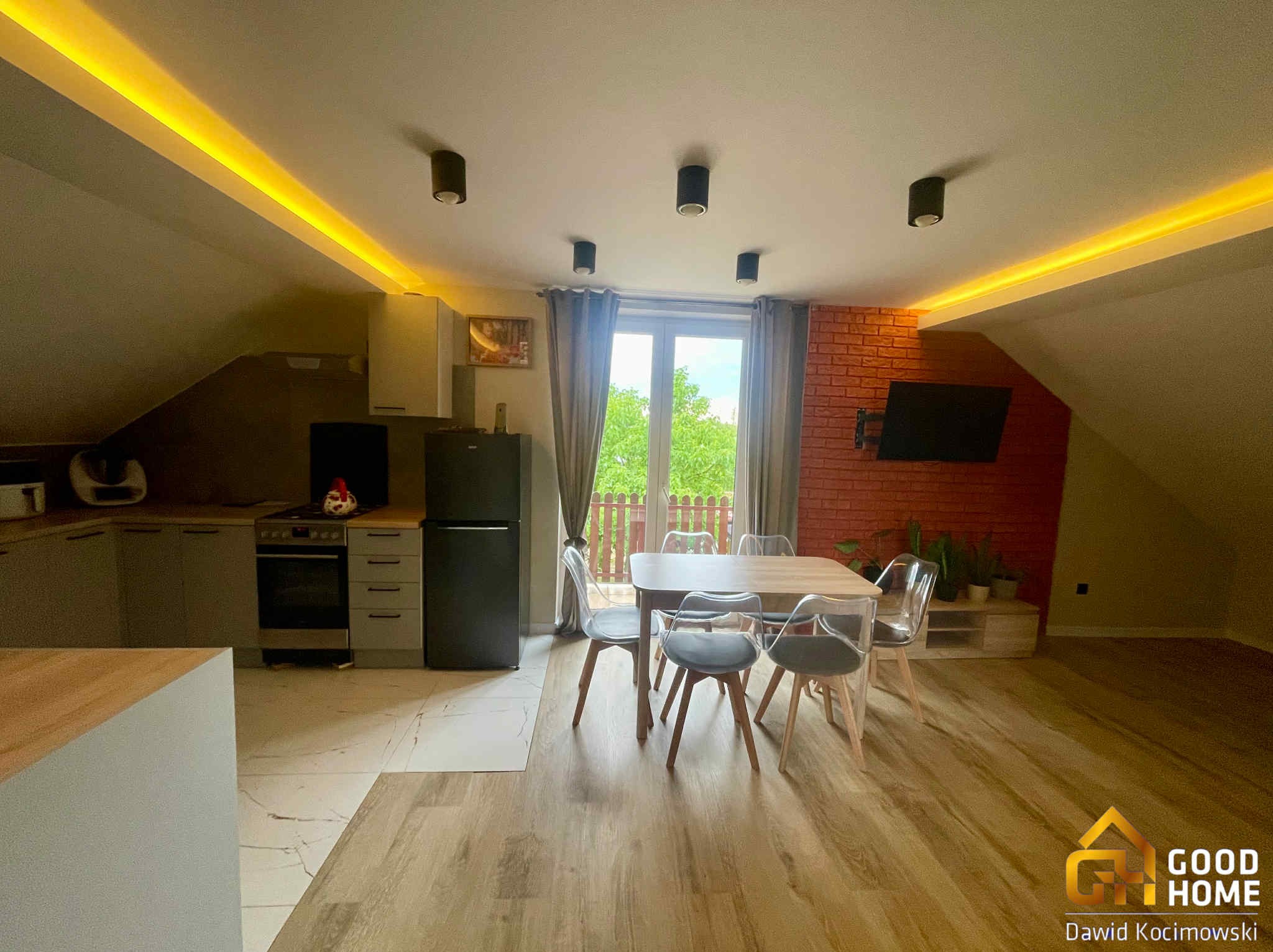 Jasne, przestronne poddasze po remoncie z aneksem kuchennym, stołem i krzesłami. Ceglana ściana z telewizorem, oświetlenie LED. Widok na zieleń za oknem.