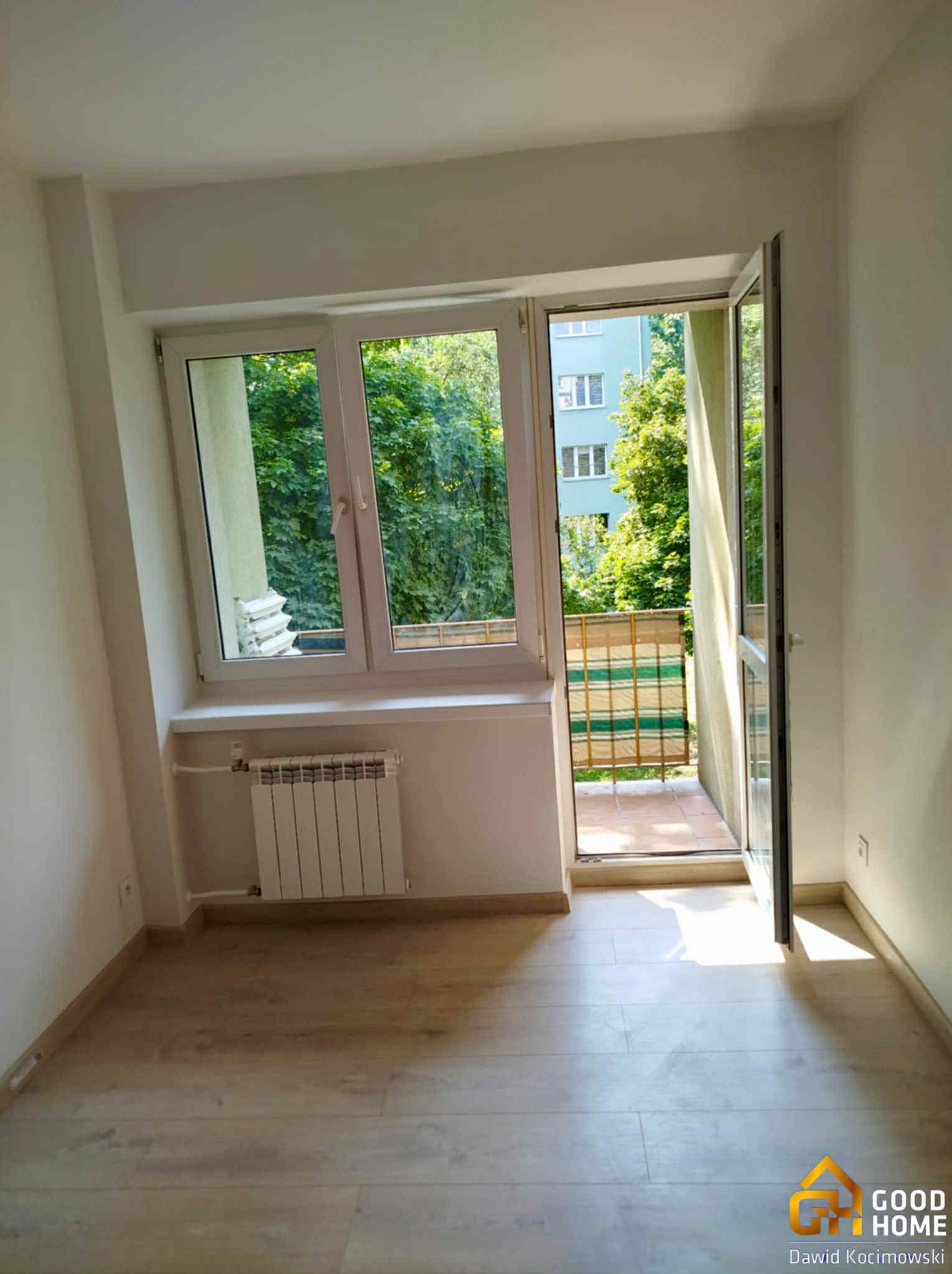 Jasne wnętrze pokoju z parkietem, oknem i otwartym balkonem. Widok na zieleń za oknem. Grzejnik pod oknem. Świeżo wykończone ściany.