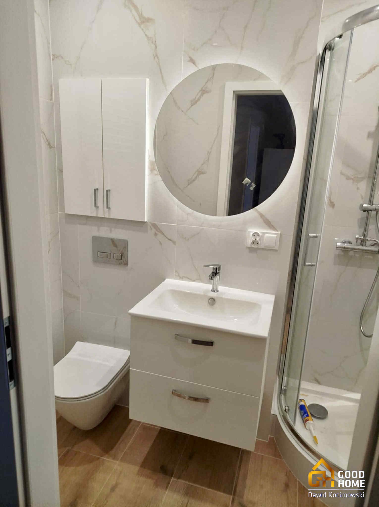 Wykończona łazienka z kabiną prysznicową, toaletą i umywalką z szafką. Białe płytki imitujące marmur, okrągłe lustro z podświetleniem LED. Drewnopodobna podłoga.