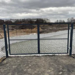 Dwuskrzydłowa brama z metalowej siatki na posesji, z widocznymi śladami niedawnych prac ziemnych i nasypem w tle, sfotografowana w pochmurny dzień.