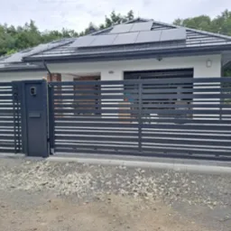 Nowoczesna, pozioma brama przesuwna z domofonem, w kolorze antracytowym, na tle domu z panelami słonecznymi i siatki ogrodzeniowej.