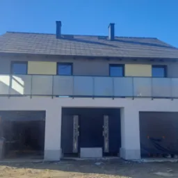 Dwukondygnacyjny dom z balkonem zabezpieczonym szklaną balustradą. Widoczne dwa garaże, drzwi wejściowe i fragment rusztowania z lewej strony.