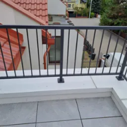 Nowoczesna, grafitowa balustrada balkonowa z pionowymi szczeblami na tle miejskiego krajobrazu z czerwonymi dachami i zielenią. Widok z góry na szare płytki balkonu.