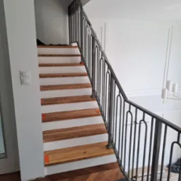 Schody z drewnianymi stopniami i czarną, metalową balustradą o dekoracyjnym wzorze. Widok z dołu schodów, na podłodze zabezpieczenia.