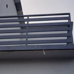 Nowoczesna, pozioma balustrada balkonowa w kolorze grafitowym, zamontowana na elewacji budynku. Widoczne detale wykończenia balkonu i fragment okna na górze.