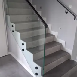 Nowoczesne schody z szarą okładziną, zabezpieczone szklaną balustradą z czarnymi mocowaniami i poręczą. Minimalistyczny design, jasne wnętrze.