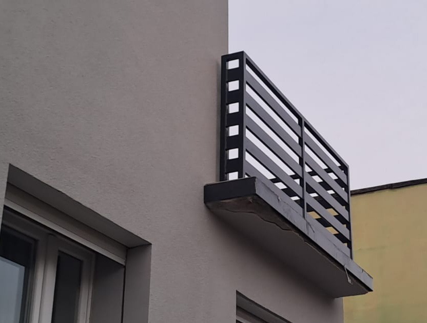 Nowoczesna, czarna balustrada balkonowa z poziomych profili, zamontowana na szarej elewacji budynku. Minimalistyczny design, widok z dołu.