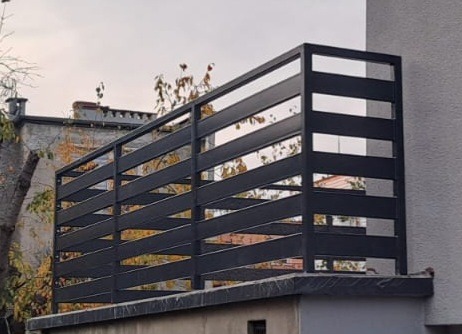 Nowoczesna, pozioma, czarna balustrada metalowa na balkonie. Minimalistyczny design z prostymi liniami. Tło stanowią budynki i drzewa w jesiennej scenerii.