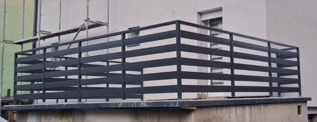 Nowoczesna, pozioma, metalowa balustrada balkonowa w kolorze antracytowym. Widoczne rusztowania w tle sugerują trwające prace wykończeniowe.