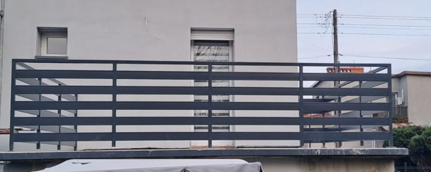 Nowoczesna, pozioma, metalowa balustrada balkonowa w kolorze grafitowym, z widocznymi oknami budynku w tle. Minimalistyczny design, proste linie.