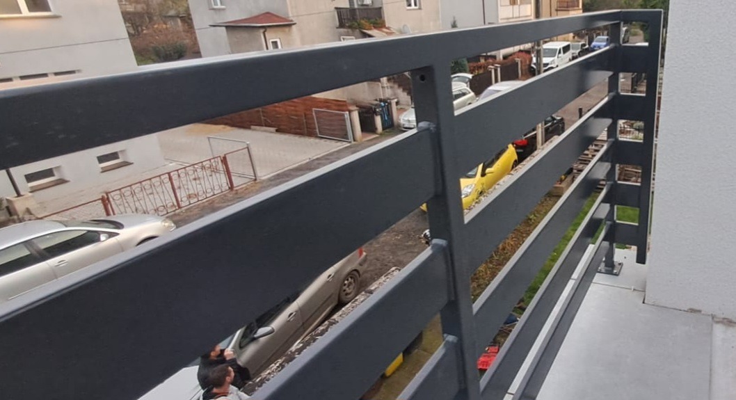 Szara, metalowa balustrada balkonowa z widokiem na ulicę i zaparkowane samochody. Prosta konstrukcja, nowoczesny design. Widok z góry, lekki kąt.